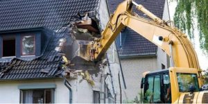 A large excavator with a steel claw demolishing a house, with bricks and debris scattered around as the walls come tumbling down.